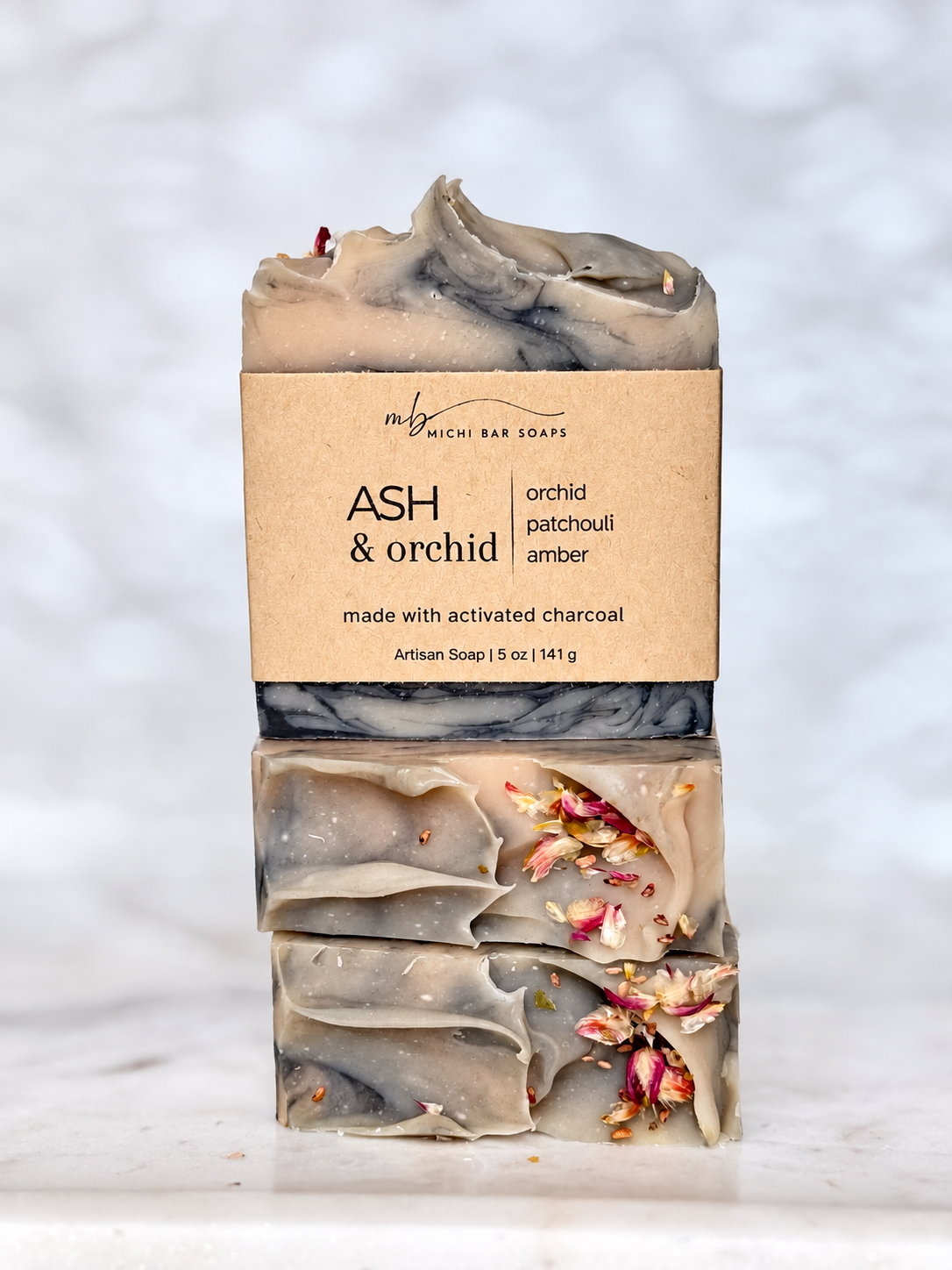 A handmade artisan soap bar labeled 'ASH & orchid' by Michi Bar Soaps, featuring a marbled gray and beige body with dried orchid petals, set against a soft-focus white marble background.