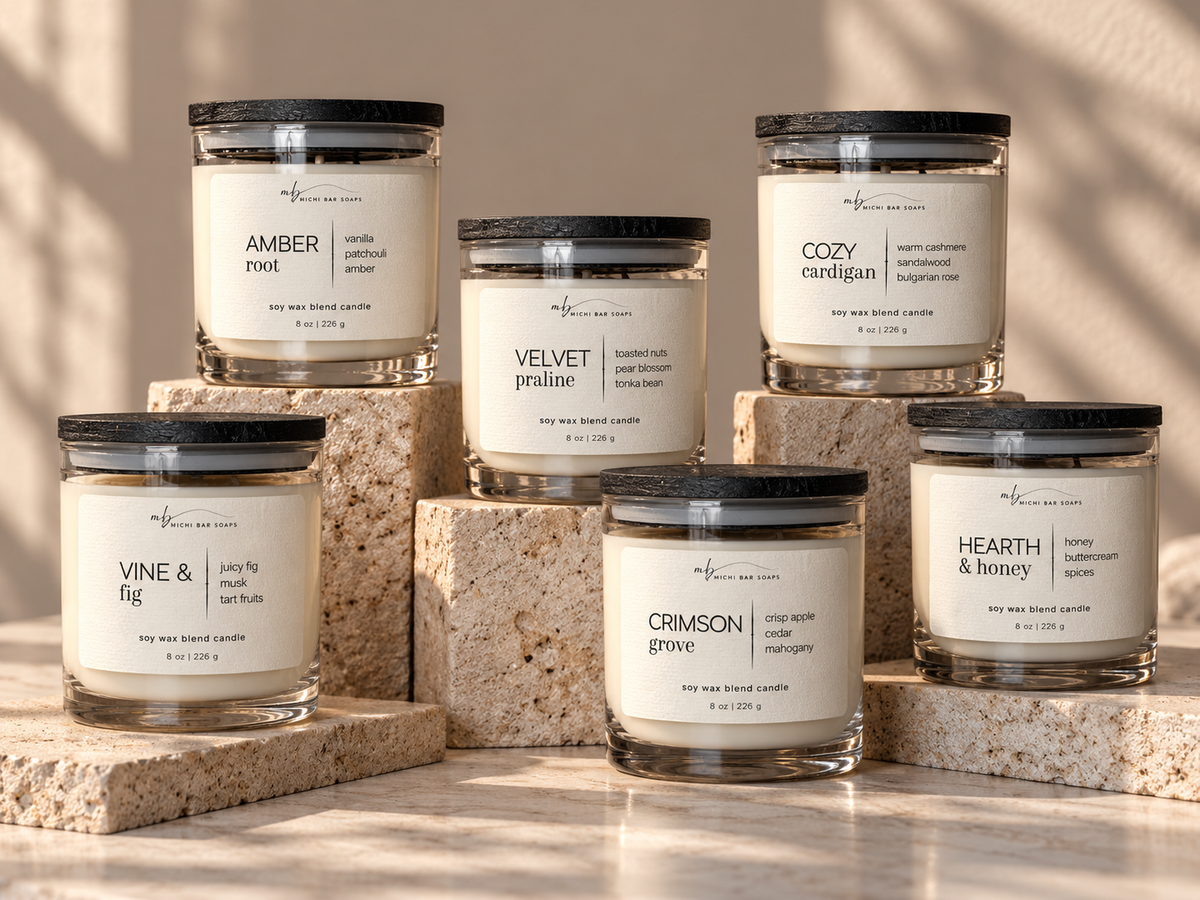 Six soy wax candle jars with black lids and cream labels are arranged on stacked stone blocks against a warm beige background. The labels show different scents: Amber Root, Velvet Praline, Cozy Cardigan, Vine & Fig, Crimson Grove, and Hearth & Honey.