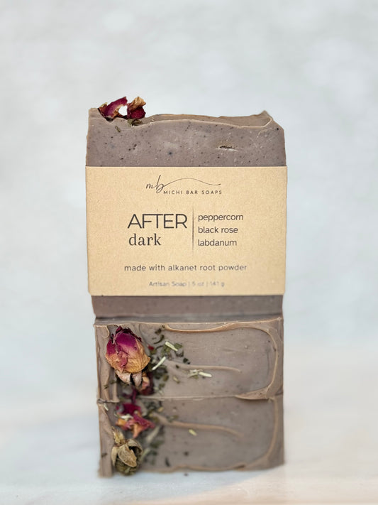 A handmade bar of soap from Michi Bar Soaps, labeled 'AFTER dark' with notes of peppercorn, black rose, and labdanum. The soap is a muted taupe-brown color with dried rose petals and herbs embedded in it, wrapped with its paper label attached.