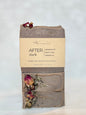 A handmade bar of soap from Michi Bar Soaps, labeled 'AFTER dark' with notes of peppercorn, black rose, and labdanum. The soap is a muted taupe-brown color with dried rose petals and herbs embedded in it, wrapped with its paper label attached.
