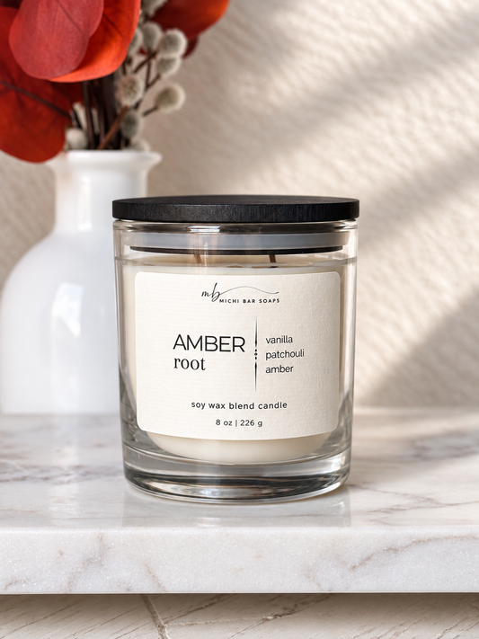 A glass jar candle with a black lid and a cream label reading 'AMBER root' and 'vanilla patchouli amber' sits on a white marble surface. A white vase with red flowers is blurred in the background against a textured beige wall with soft shadows.