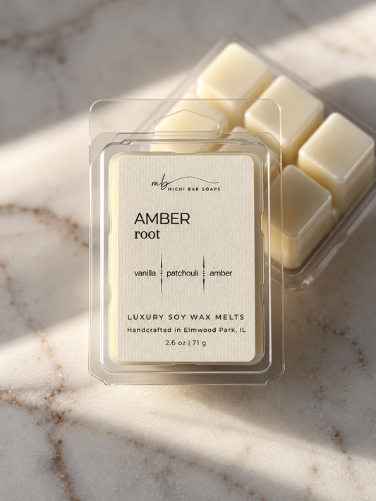 A clear clamshell package of Michi Bar Soaps 'Amber Root' luxury soy wax melts sits on a light marble surface with soft diagonal shadows. A second package showing cream-colored wax cubes is partially visible behind it.