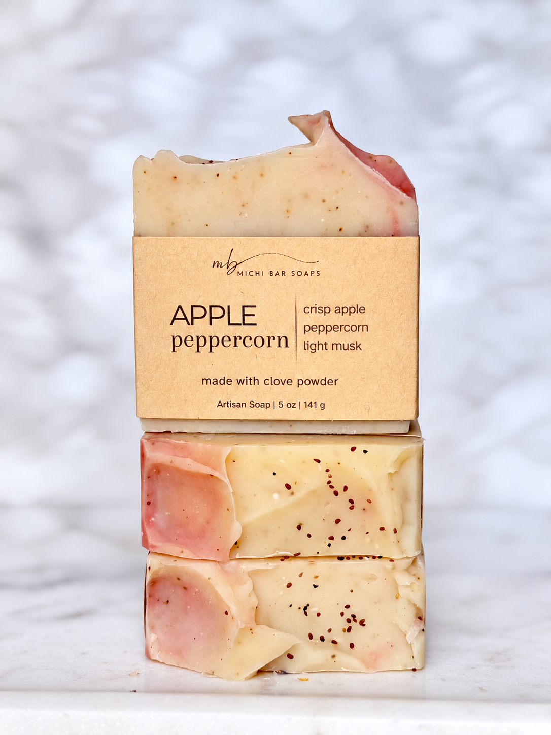 Three stacked bars of Michi Bar Soaps 'Apple Peppercorn' artisan soap, showing the cream-colored bars with pinkish-red edges and dark pepper flakes, wrapped in a kraft paper band against a white marble background.
