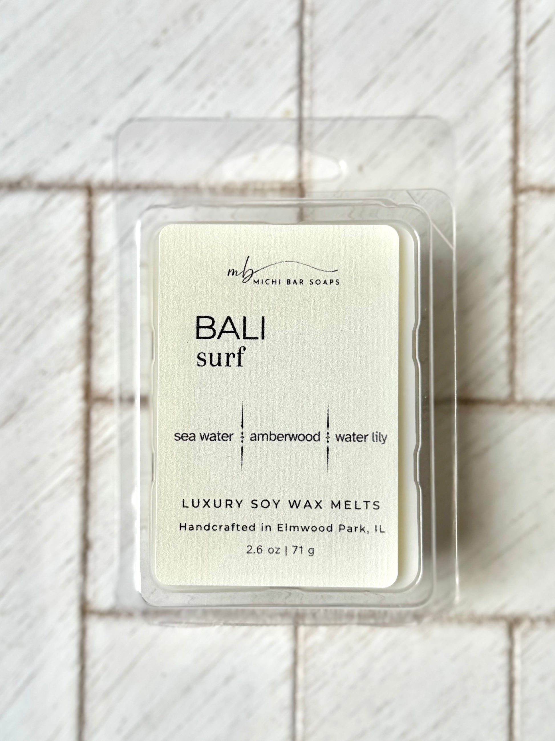 clear clamshell mold with wax scented. cream label that says Bali Surf and scent notes of sea water, amberwood and water lily