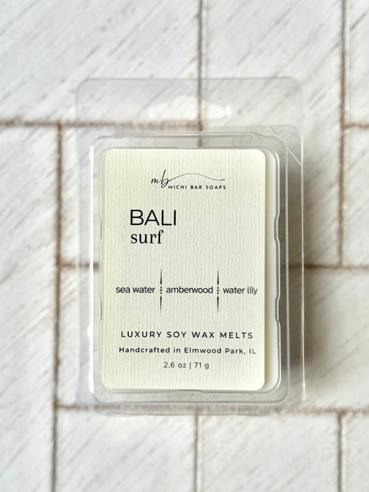 clear clamshell mold with wax scented. cream label that says Bali Surf and scent notes of sea water, amberwood and water lily