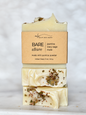 Three stacked bars of Michi Bar Soaps 'Bare Allure' artisan soap on a light marble surface with a soft, out-of-focus white background. The soap is creamy off-white with dried botanicals and is wrapped in a kraft paper label.