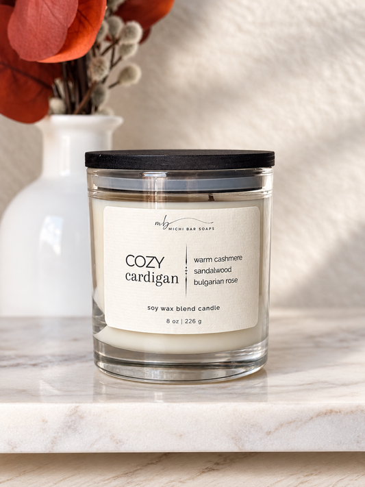 A glass jar candle with a black lid and a cream label reading 'COZY cardigan' sits on a light marble surface. A white vase with red leaves is blurred in the background against a soft beige wall.