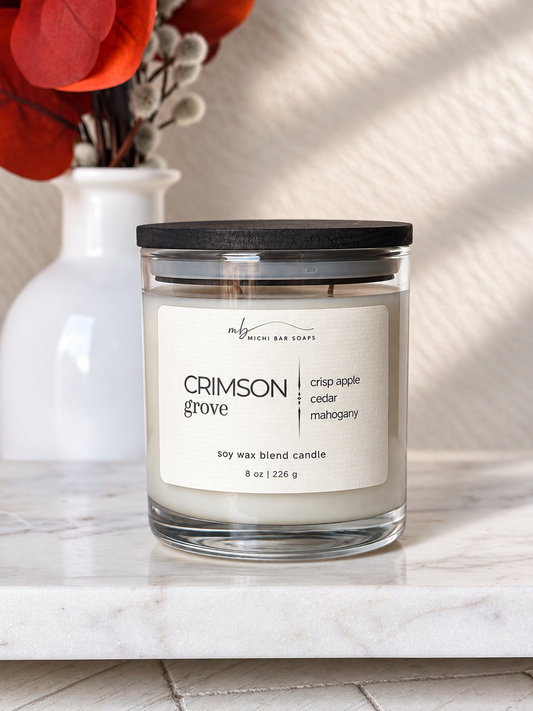 An 8 oz soy wax blend candle by Michi Bar Soaps in a clear glass jar with a black lid and a cream label reading 'CRIMSON grove' and 'crisp apple cedar mahogany.' The candle sits on a white marble surface with a white ceramic vase and red dried leaves in the background.