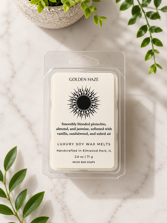 A clear clamshell package of Golden Haze luxury soy wax melts sits on a light marble surface with green leaves and a small plant around it. The front label is cream with black text and a sunburst logo.