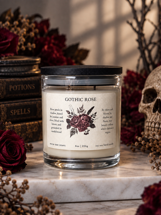 A glass jar candle labeled 'Gothic Rose' by Michi Bar Soaps, styled on a marble surface with dark red roses, dried botanicals, a skull, and vintage-style books in the background.