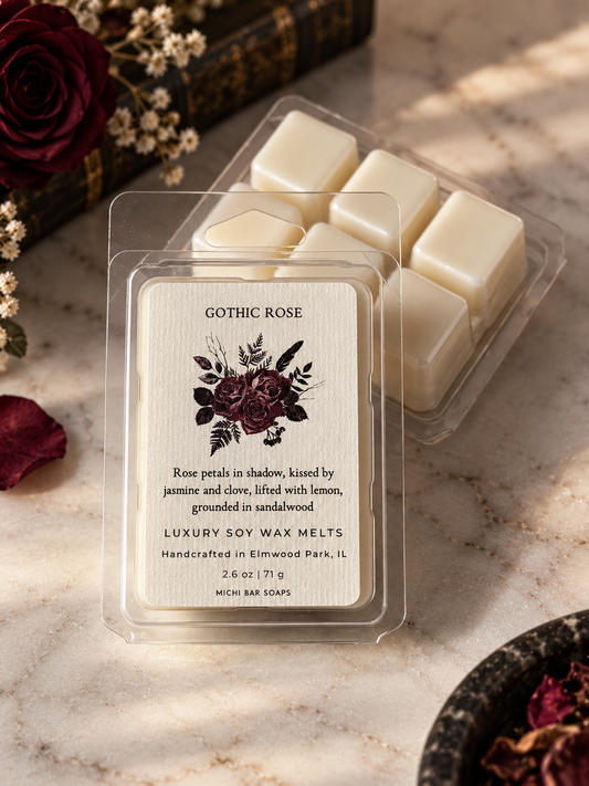 A clear plastic clamshell package of Gothic Rose Luxury Soy Wax Melts sits on a marble surface, with additional cream-colored wax melts visible behind it. The front label reads 'Gothic Rose' with floral artwork and scent notes, and the brand 'Michi Bar Soaps' appears at the bottom.