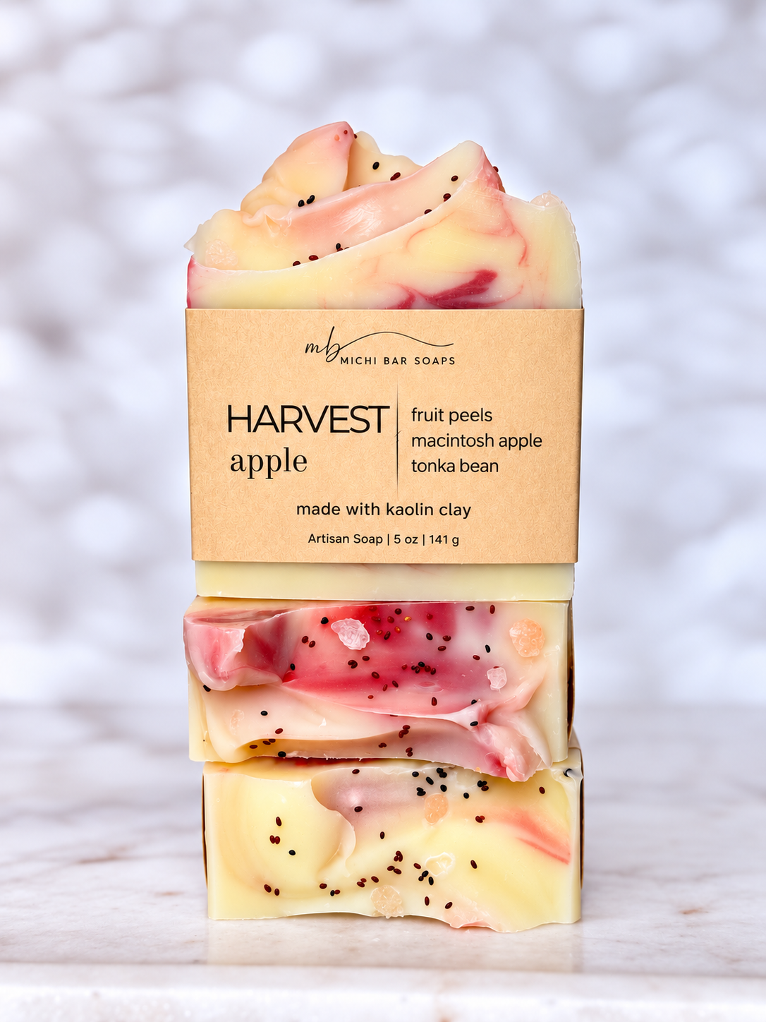 Three stacked artisanal bar soaps from Michi Bar Soaps in a 'Harvest Apple' scent, wrapped in a kraft paper label, set on a white marble surface with a soft marble background.