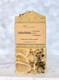 A handmade bar of soap labeled 'HAVANA' by Michi Bar Soaps, made with turmeric powder. The soap is a pale, creamy beige color with visible dried green and brown herbs embedded within, shown on a light marble surface against a white marble background.