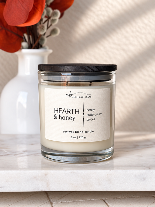 A glass jar candle with a black lid and a cream label reading 'HEARTH & honey' sits on a white marble surface. A white vase with red-orange dried leaves is blurred in the background against a textured beige wall with soft shadows.