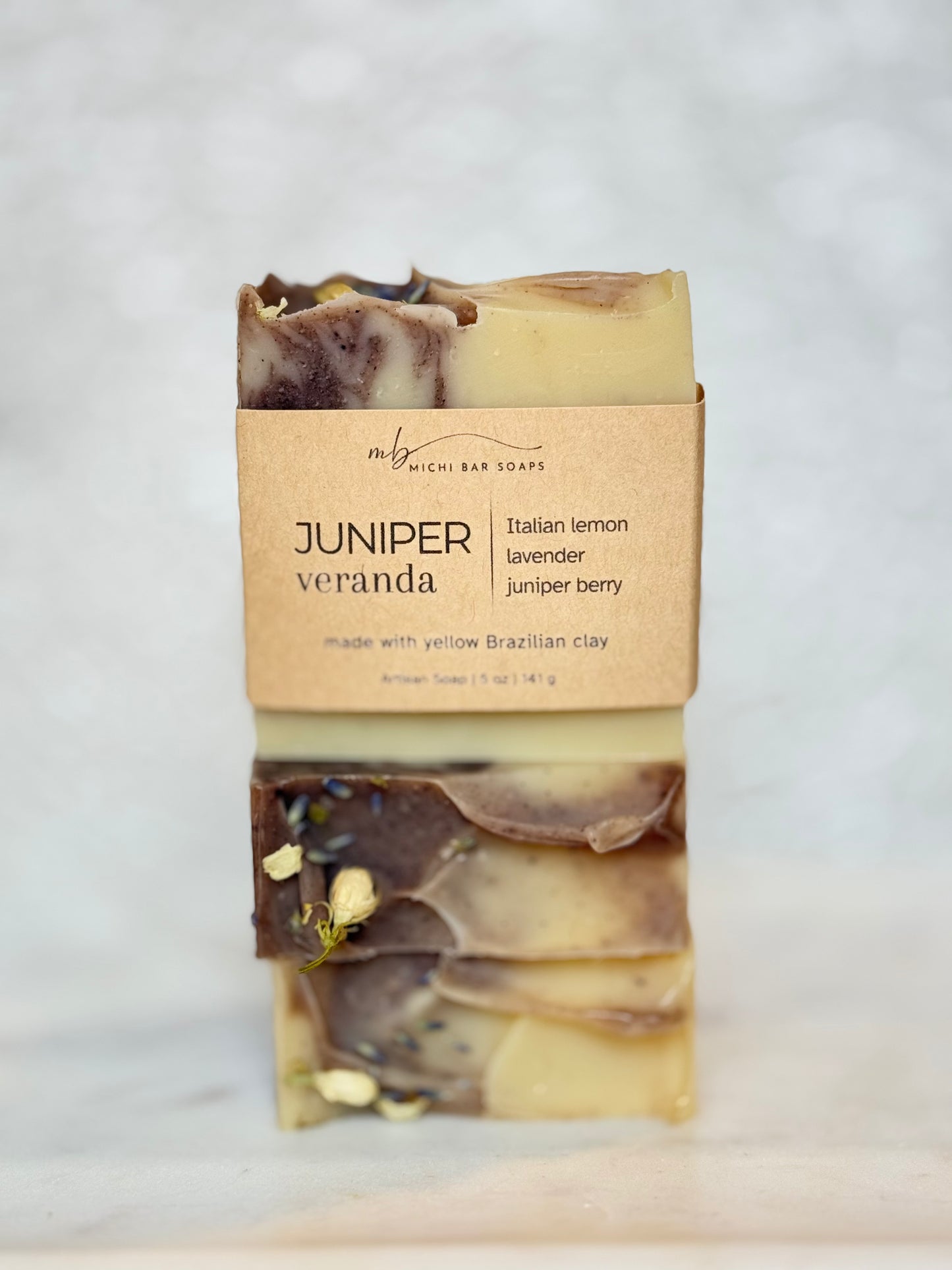 A stacked bar of handmade soap labeled 'JUNIPER veranda' by My Mighty Bar Soaps, wrapped in a kraft paper band, set against a light marble background.