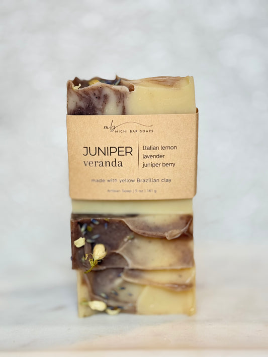 A stacked bar of handmade soap labeled 'JUNIPER veranda' by My Mighty Bar Soaps, wrapped in a kraft paper band, set against a light marble background.