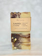 A stacked bar of handmade soap labeled 'JUNIPER veranda' by My Mighty Bar Soaps, wrapped in a kraft paper band, set against a light marble background.
