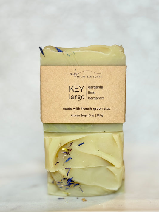 A bar of handmade soap labeled 'KEY largo' by Rich Bar Soaps, featuring a light yellow-green base with blue dried flower accents, wrapped in a kraft paper label on a light marble surface.