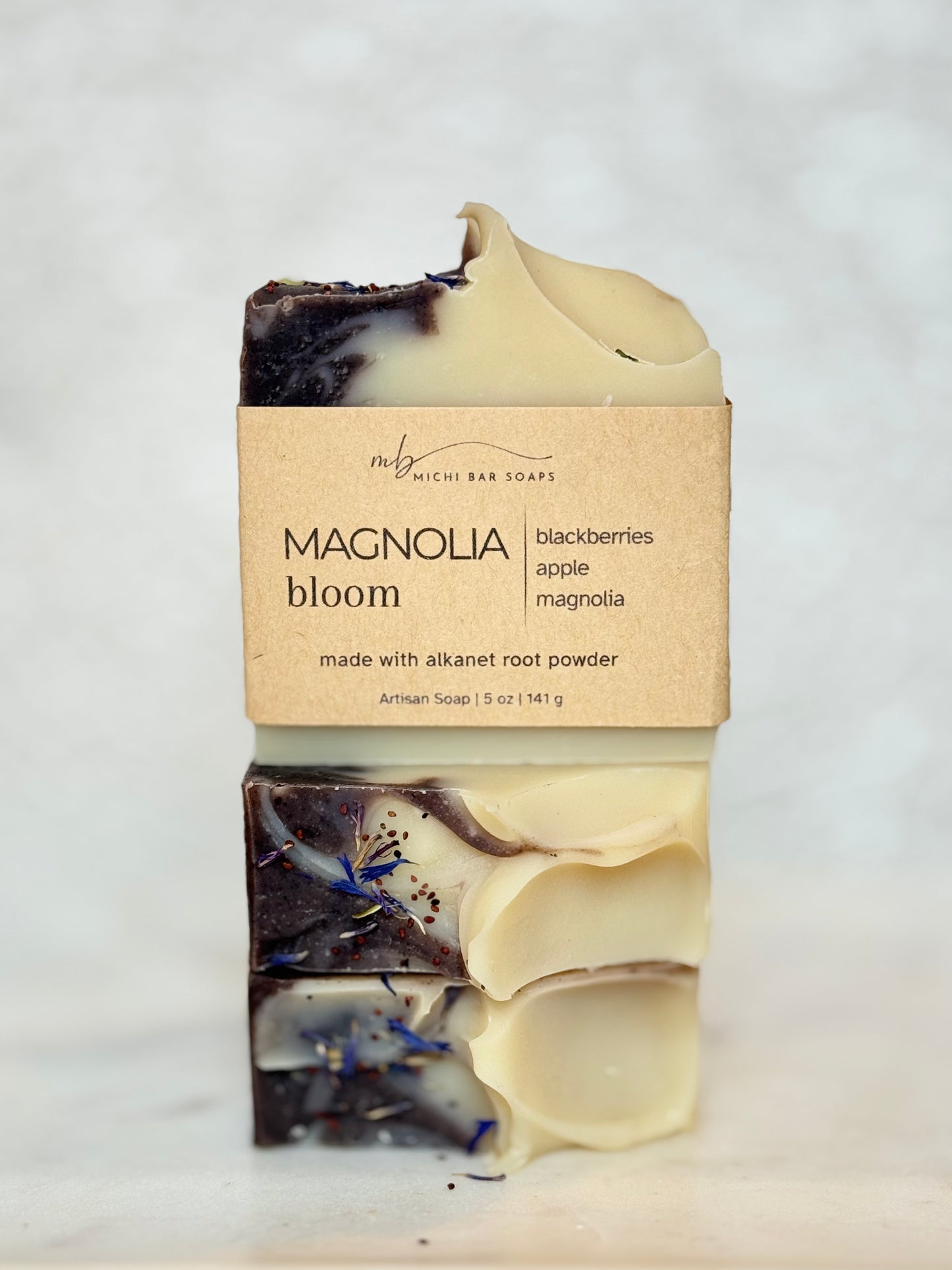 A handmade artisan soap bar wrapped in a kraft paper label that reads 'MAGNOLIA bloom' and 'blackberries apple magnolia made with alkanet root powder' by 'The M' Michi Bar Soaps, set against a light marble background.
