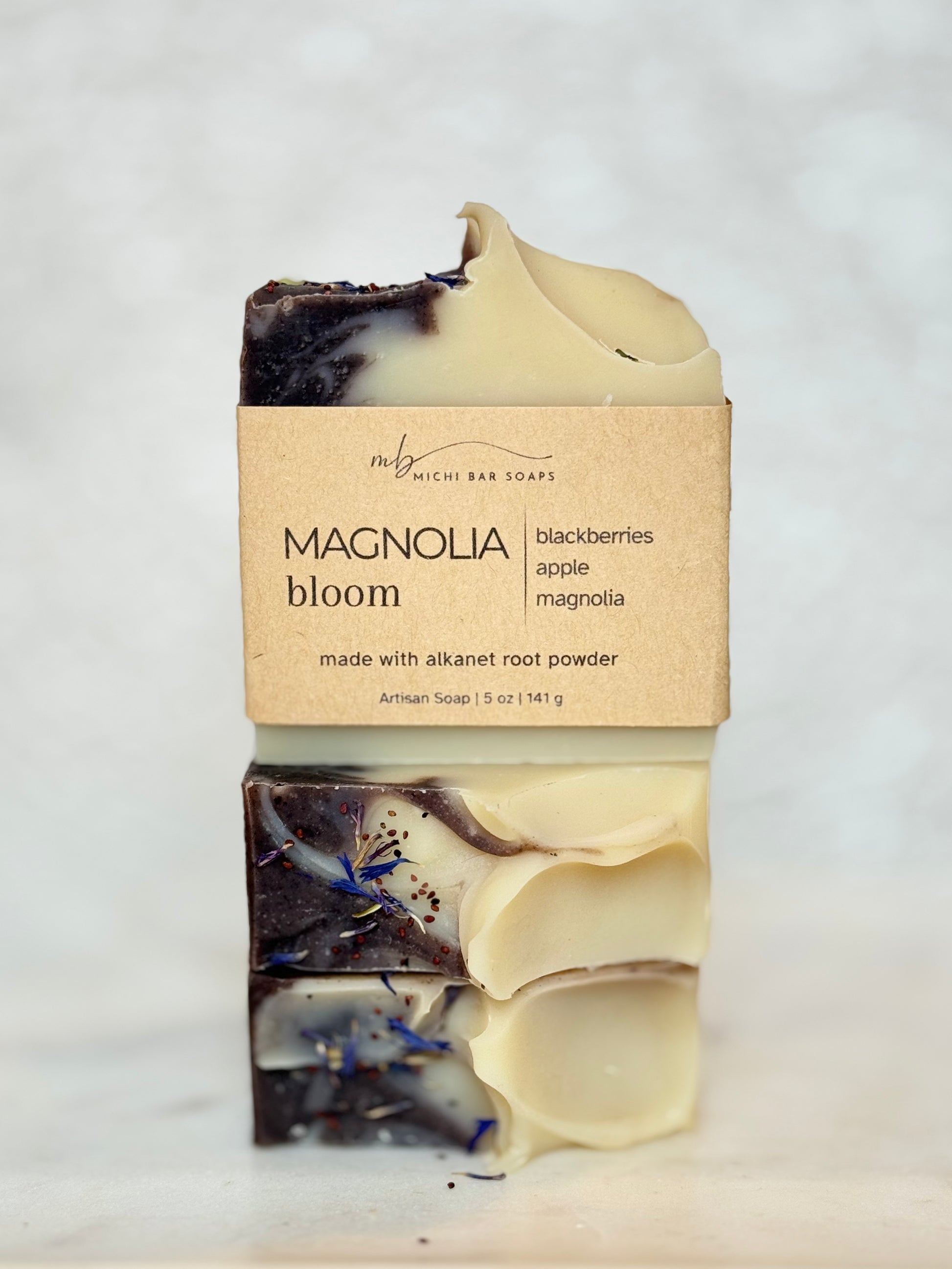 A handmade artisan soap bar wrapped in a kraft paper label that reads 'MAGNOLIA bloom' and 'blackberries apple magnolia made with alkanet root powder' by 'The M' Michi Bar Soaps, set against a light marble background.