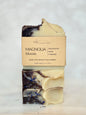 A handmade artisan soap bar wrapped in a kraft paper label that reads 'MAGNOLIA bloom' and 'blackberries apple magnolia made with alkanet root powder' by 'The M' Michi Bar Soaps, set against a light marble background.
