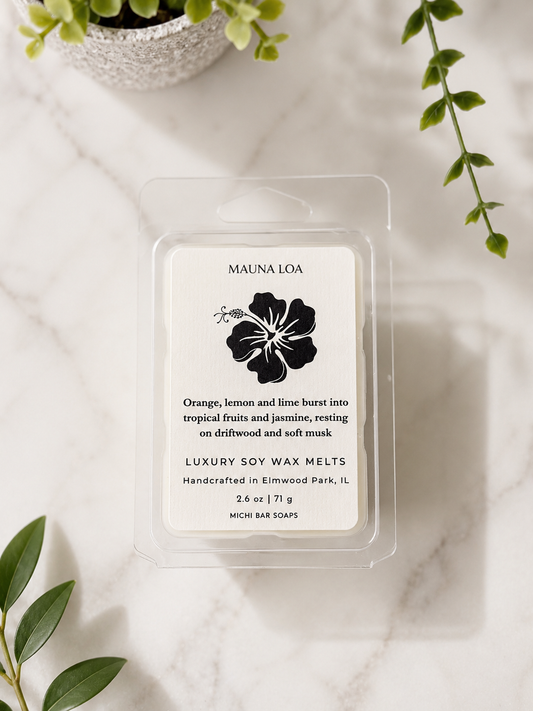 A clear plastic clamshell package of Mauna Loa luxury soy wax melts sits on a white marble surface with green leaves and a small plant in the background. The label is white with black text and a black hibiscus flower illustration.