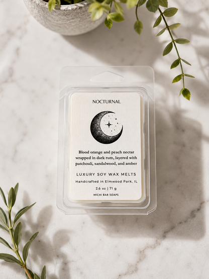 A clear plastic clamshell package of Nocturnal soy wax melts sits on a light marble surface with green leaves and a small plant in the background. The label reads 'NOCTURNAL' and 'Blood orange and peach nectar wrapped in dish rum, layered with patchouli, sandalwood, and amber.