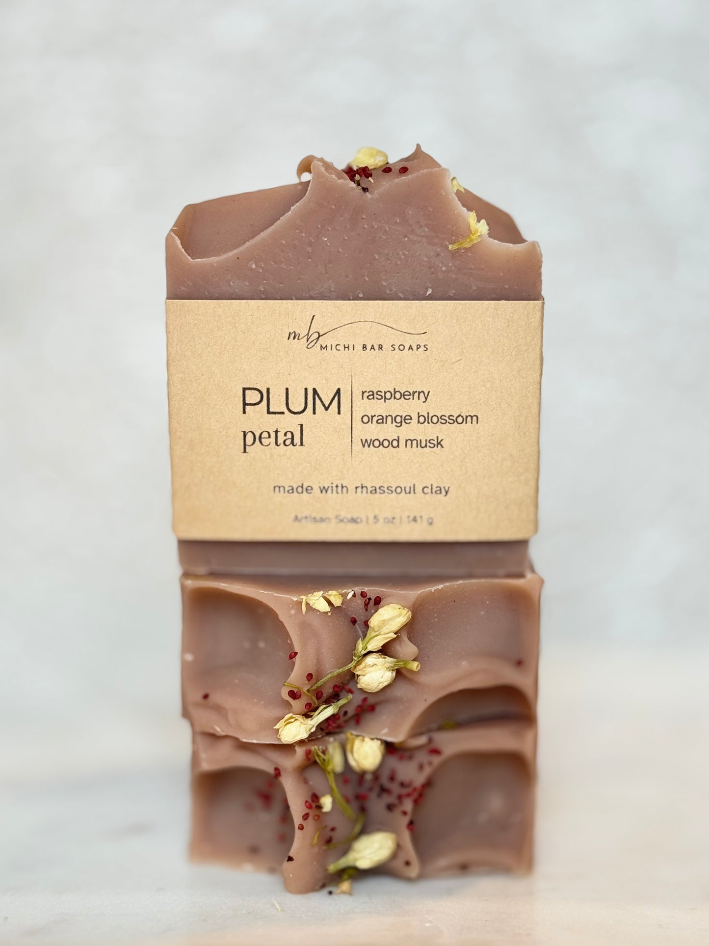 A stacked bar of Rich Bar Soaps 'Plum Petal' artisan soap, showing the top and bottom halves with dried petals and a kraft paper label, set against a light marble background.