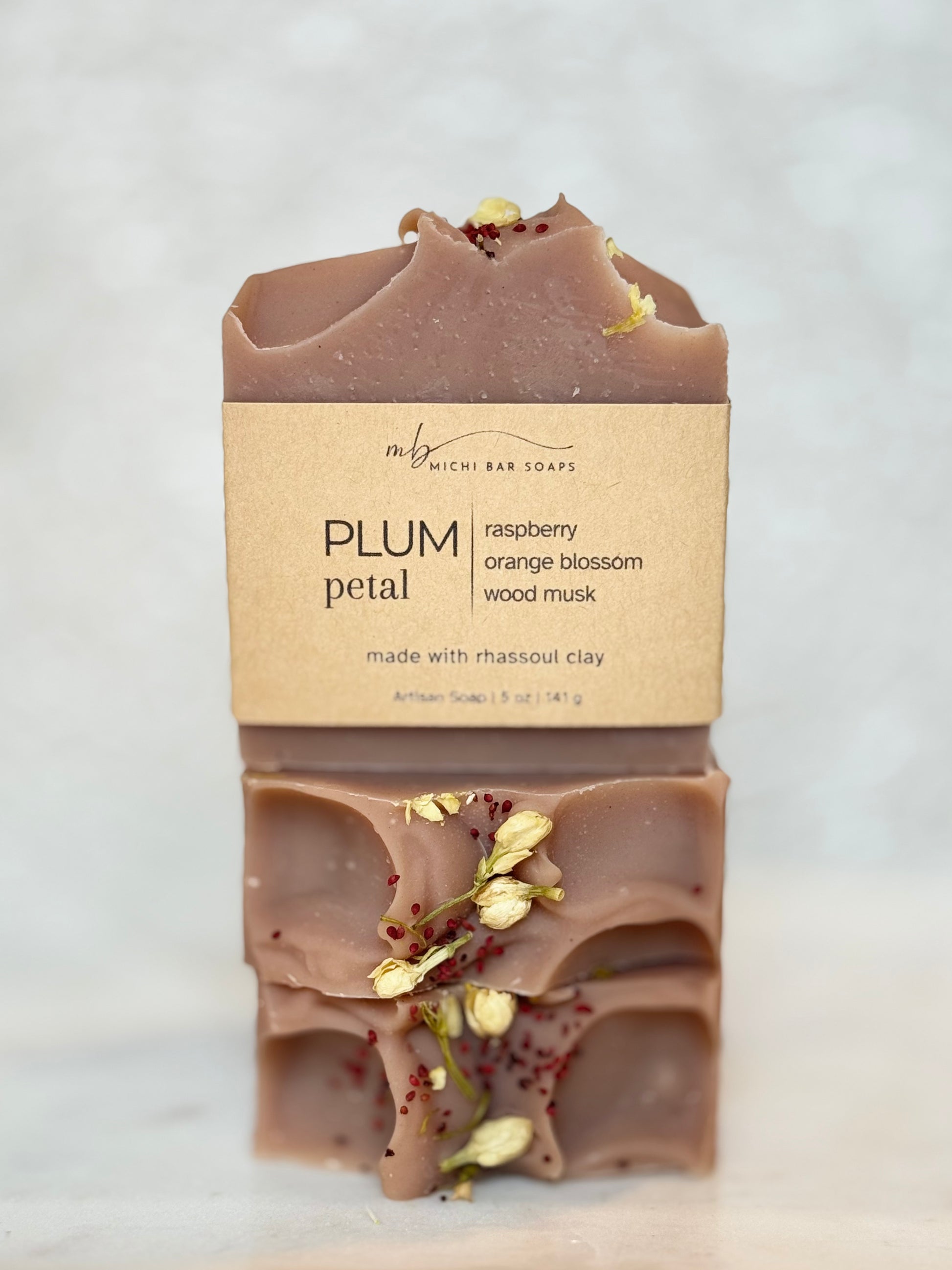 A stacked bar of Rich Bar Soaps 'Plum Petal' artisan soap, showing the top and bottom halves with dried petals and a kraft paper label, set against a light marble background.