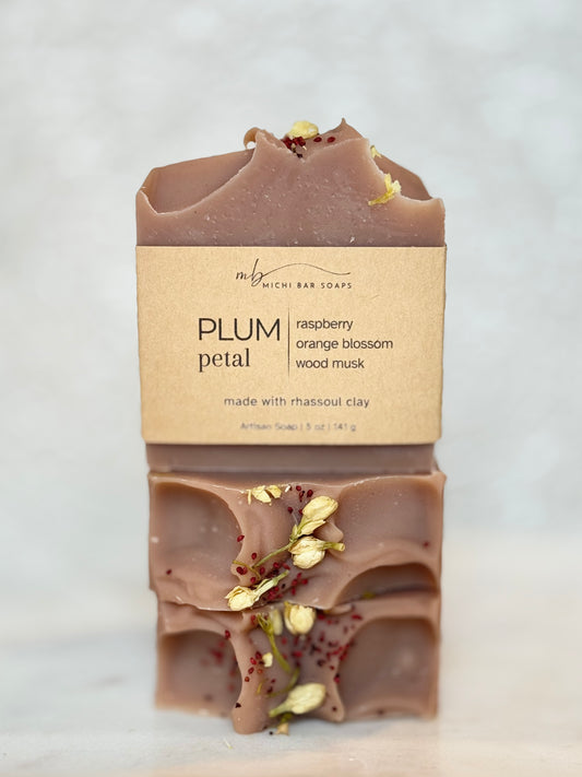 A stacked bar of Rich Bar Soaps 'Plum Petal' artisan soap, showing the top and bottom halves with dried petals and a kraft paper label, set against a light marble background.