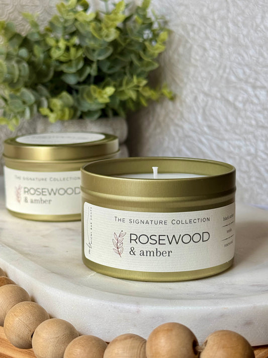 Two 'Rosewood & Amber' candles in gold tins on a marble surface with a plant in the background.