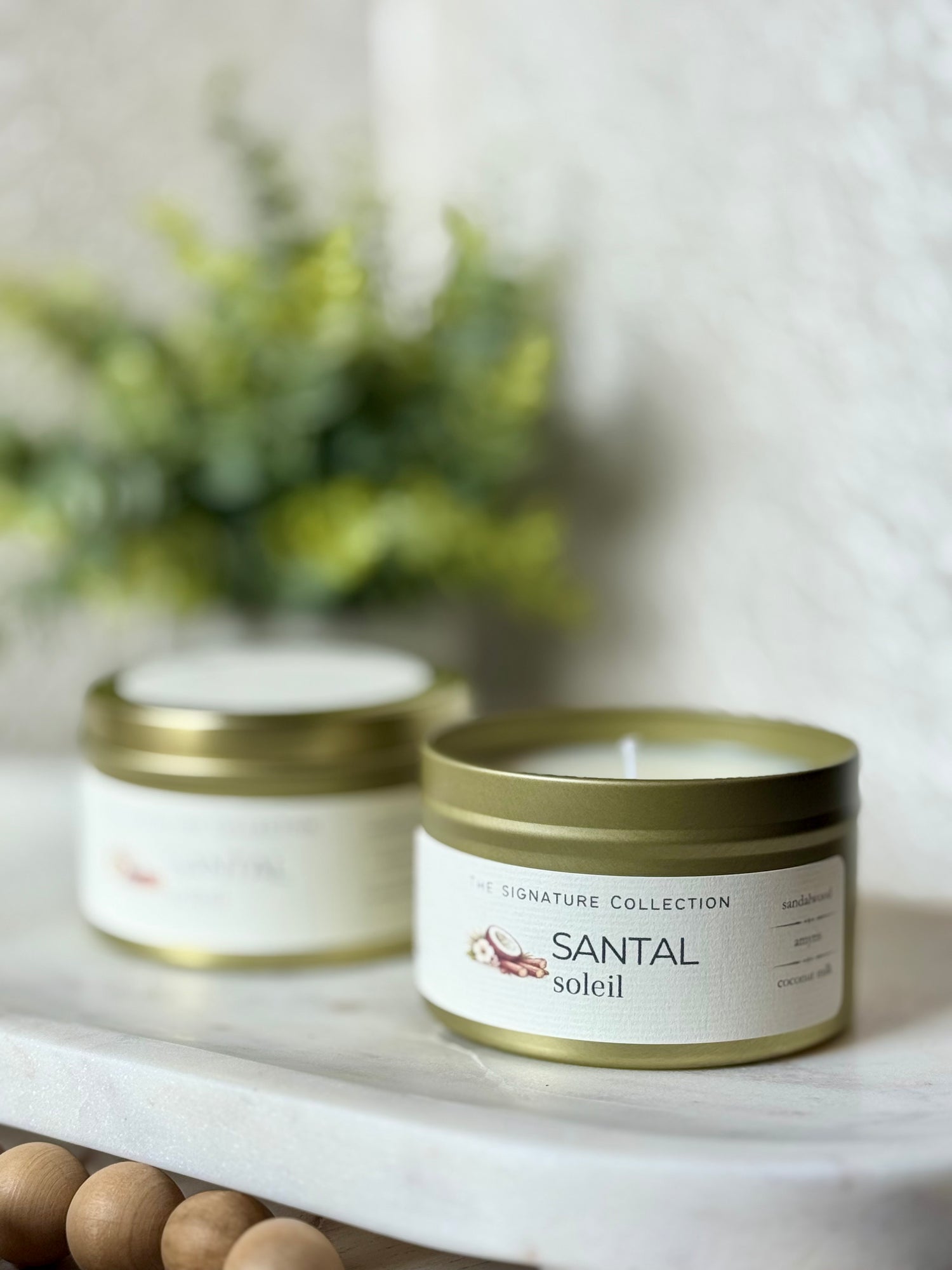 Two Santal Soleil candles on a marble surface with a blurred plant in the background.