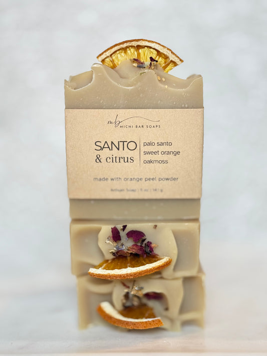 A handmade soap bar package labeled 'SANTO & CITRUS' by Milch Bar Soaps, with dried orange slices and purple flower petals embedded in pale cream soap, photographed on a soft light gray background.