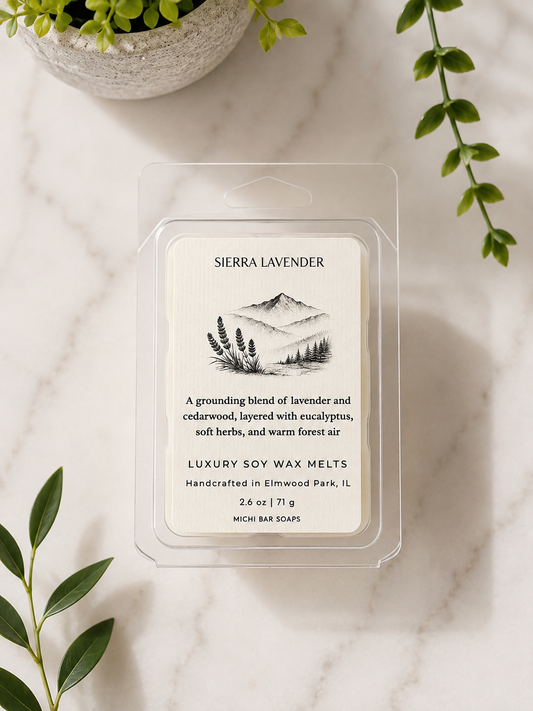 A clear plastic clamshell package of Sierra Lavender luxury soy wax melts sits on a light marble surface with green leaves and a small plant pot as styled props. The label is cream with black text and a mountain-and-plant illustration.