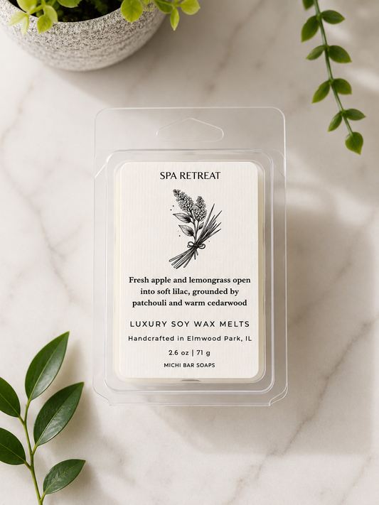 A clear plastic clamshell package containing a single wax melt labeled 'Spa Retreat' by Michi Bar Soaps, placed on a light marble surface with green leaves and a small plant in the background.