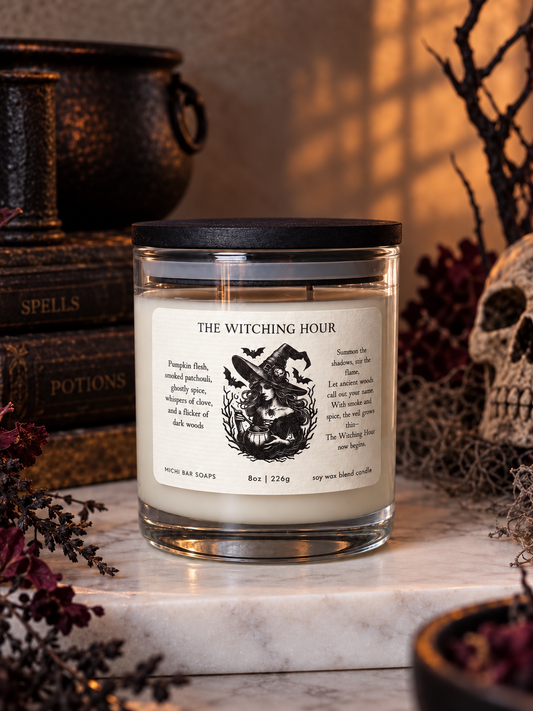 A glass jar candle with a black lid and a cream label reading 'THE WITCHING HOUR' sits on a marble surface surrounded by Halloween-themed props including a skull, dried flowers, and old books.