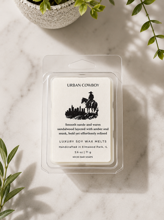 A packaged wax melt product labeled Urban Cowboy sits on a light marble surface with green leaves and a plant pot around it. The clear plastic clamshell contains a white label with black text and a black silhouette illustration.