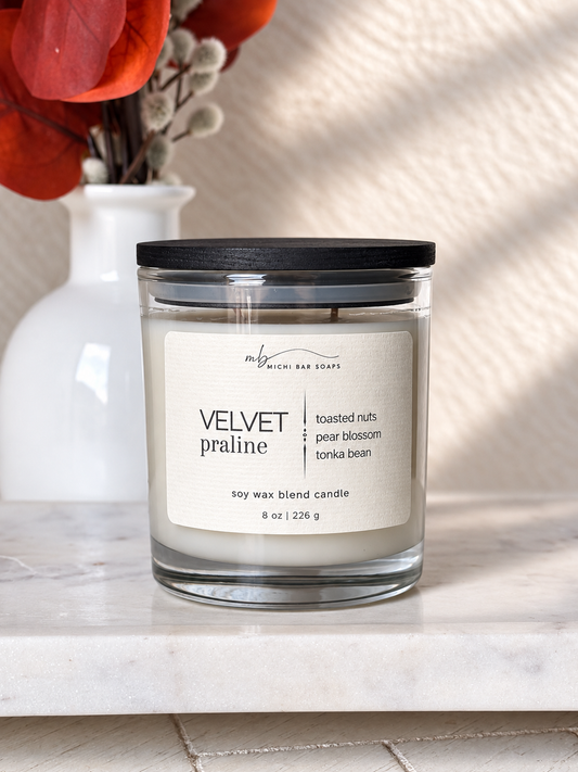 A jar candle labeled 'VELVET pralinee' by Michi Bar Soaps sits on a light marble surface. The candle is in a clear glass jar with white wax and a black lid, with a white vase and red dried leaves blurred in the background.
