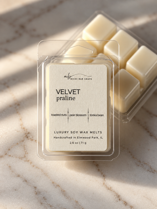A clear clamshell package of Velvet Praline luxury soy wax melts sits on a light marble surface with a second tray of wax melts behind it. The wax melts are creamy off-white, and the packaging features a textured cream label with black text.