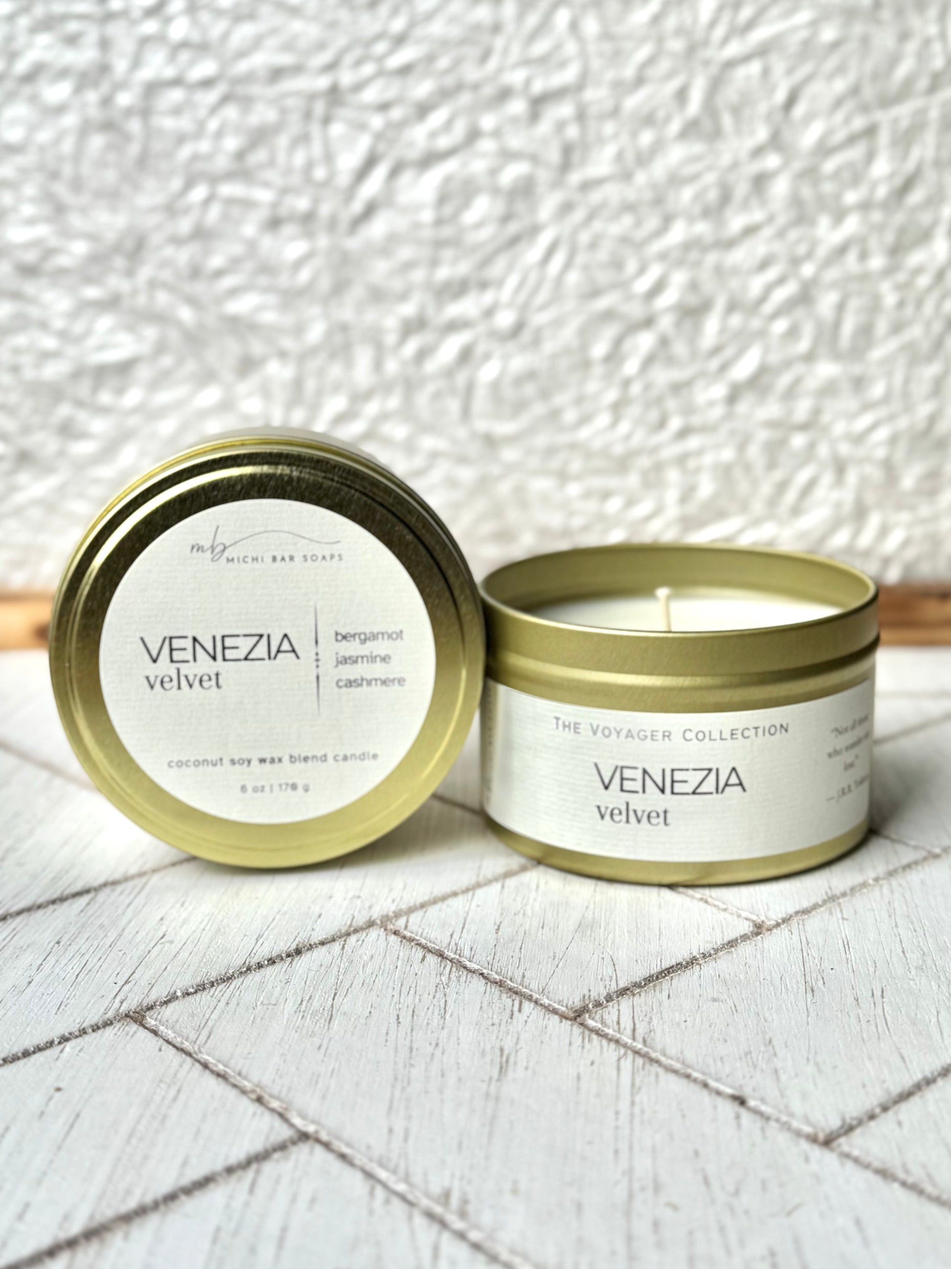 A gold-colored tin candle with the label 'VENEZIA VELVET' on a white textured surface, with a lit candle inside the tin, placed on a white tiled surface.
