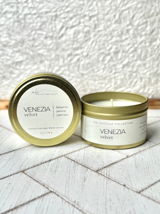 A gold-colored tin candle with the label 'VENEZIA VELVET' on a white textured surface, with a lit candle inside the tin, placed on a white tiled surface.