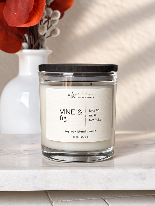 A soy wax blend candle in a clear glass jar with a black lid and a white label reading 'VINE & fig' and 'michi bar soaps', set on a white marble surface with a white vase and red-orange leaves blurred in the background.