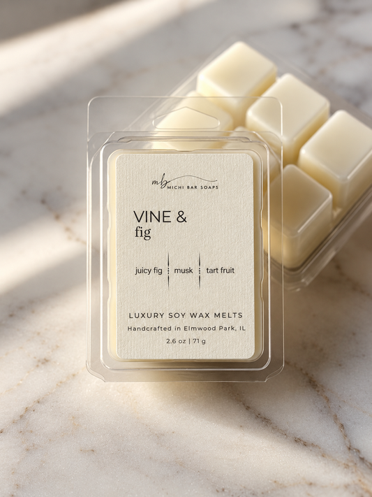 A clear clamshell package of Vine & Fig luxury soy wax melts sits on a light marble surface, with additional wax melts visible behind it. The label reads 'VINE & fig' and 'LUXURY SOY WAX MELTS' with scent notes 'juicy fig | musk | tart fruit.