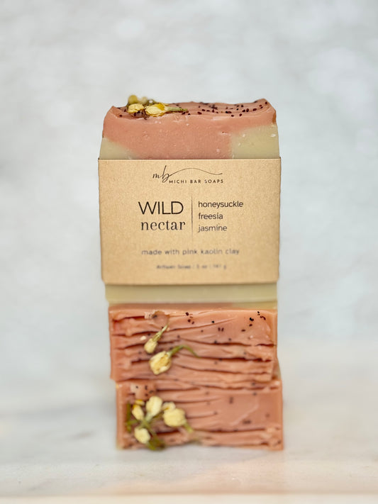 A single bar of soap from the brand 'my precious bar soaps' named 'WILD nectar', wrapped in a kraft paper sleeve, standing vertically on a light marble surface against a white marble background.